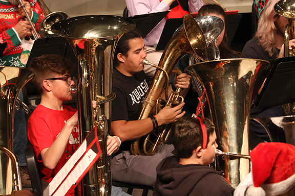 Several tuba players in holiday attire playing Christmas music on their instruments on a stage.
