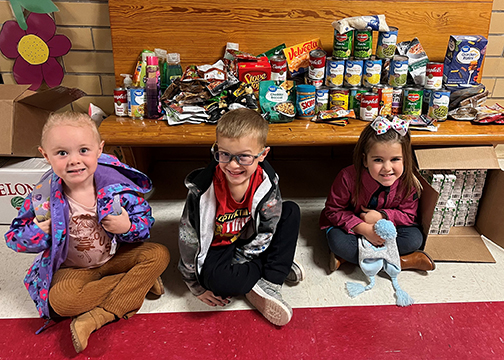 PreK and Kindergarten students sitting in front of several grocery items collected as part of a Day of Kindness project.