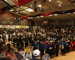 Wide-angle photograph of Northwestern Oklahoma State University's Fall Commencement ceremony in Percefull Fieldhouse. Graduates in black caps and gowns are seated in rows in the center of the gymnasium floor, with faculty in colorful academic regalia seated in front. Bleachers filled with family and friends surround the ceremony space. Red and black championship banners hang from the ceiling, and Northwestern's Ranger mascot logo is visible on the wall.