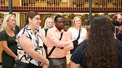 A group of adults standing in the J.W. Martin Library on the Northwestern Oklahoma State University campus, listening to a presentation. The setting shows two-story library bookshelves and warm overhead lighting in the background. In the foreground, a woman wearing glasses and a floral top stands next to a woman in an olive-green dress. A man in a peach button-up shirt stands with his arms crossed beside them. Two women in light-colored tops and a man in a Northwestern Homecoming shirt are visible in the background. The group's attention is directed toward a presenter whose back is to the camera. The photo was taken during the 2025 DNP Residency Week poster presentation.