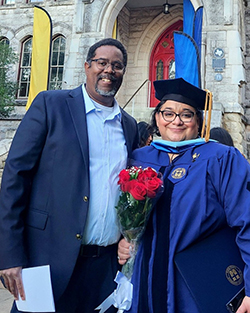 Dr. Melissa Brown in blue doctoral regalia with gold tassel and St. Edward's University insignia holding red roses standing with her husband Dr. Roger Brown in dark suit outside historic stone building with red arched doorway and decorative banners at St. Edward's University graduation ceremony in Austin, Texas.