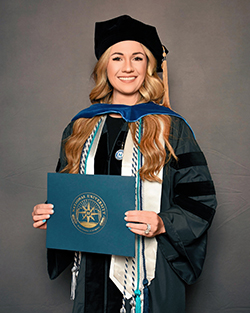 Dr. Bailey Lancaster in black doctoral tam and velvet gown with blue hood and National University seal holding diploma cover with white and teal honor cords in professional graduation portrait at National University in San Diego, California