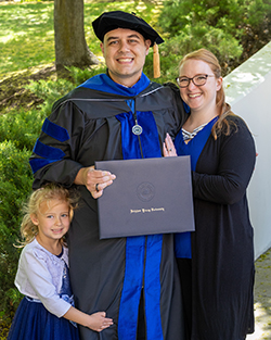 Dr. Christopher Neu in black doctoral tam and blue-trimmed regalia with Brigham Young University insignia holding diploma cover, standing outdoors with his wife and young daughter in lavender dress at graduation ceremony with trees in background at Brigham Young University.