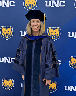 Dr. Heidi Ritchie in navy blue doctoral gown with velvet panels and gold trim wearing black doctoral tam with gold tassel standing before University of Northern Colorado Bears logo backdrop at commencement in Greeley, Colorado.