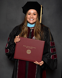Dr. Olivia Yandel in black doctoral tam and gown with turquoise and maroon hood trim holding Southern Nazarene University diploma cover in professional graduation portrait at Southern Nazarene University in Bethany.