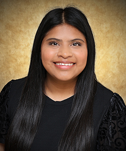 Professional headshot of Itzel Castillo, a Northwestern Oklahoma State University senior social work major from Laverne, Oklahoma. She is photographed against a warm golden-brown studio background, wearing a black top with subtle embroidered detailing, and smiling at the camera. Her long dark hair falls straight past her shoulders.