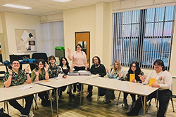 Group photo of nine NWOSU theatre students and their director seated at classroom tables in a rehearsal space. Director Lilith Fererro, wearing a tan sweater, stands in the center behind the seated cast members. Cast includes Emily Wright, Aidan Slater, Jacob Saucedo, Jaden Matthews, Bella Anguino, Matthew Skinner, and Sophie Jungman, who are displaying peace signs and smiling at the camera. Students are holding yellow script books. The classroom has white walls, vertical blinds on windows, and office furniture visible in the background.