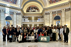 A large group of social work students and professionals from universities across Oklahoma stand together in the marble rotunda of the Oklahoma State Capitol for the 2026 Legislative Education and Advocacy (LEAD) Day in March. Several participants in the front row kneel while holding two signs: one reading 