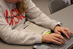 Northwestern nursing student wearing gray NWOSU sweatshirt studies from an open textbook while typing on a laptop, with a yellow highlighter visible on the desk.