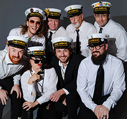 Eight members of King Cabbage Brass Band posed together against a gray studio backdrop, all wearing matching white nautical-style caps labeled "King Cabbage." Most members wear white dress shirts with dark ties; one member wears a black jacket. Members display varying facial expressions ranging from smiling to playful. One member in the front row wears dark sunglasses and holds up one finger.