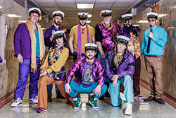 Nine members of King Cabbage Brass Band posed in a fluorescent-lit hallway wearing vibrant Mardi Gras-themed outfits in purple, gold, and teal, along with their signature white "King Cabbage" nautical caps. Outfits include sequined jackets, colorful scarves, satin blazers, and bold accessories. Two members crouch in the front row.