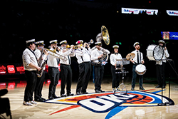 King Cabbage Brass Band performing on the Oklahoma City Thunder basketball court at center court, in front of the OKC Thunder logo. Ten musicians in white shirts and black pants play brass and percussion instruments including saxophone, trombone, trumpet, sousaphone, snare drum, cymbal, and bass drum. Bally Sports and QuikPrint signage is visible in the arena background.
