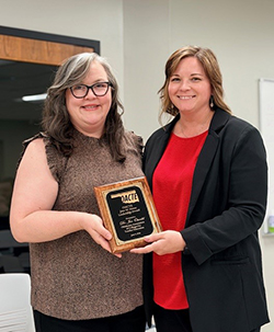 Dr. Toni Ivey, professor of education at Oklahoma State University, presents Dr. Jen Oswald, chair of Northwestern Oklahoma State University's Division of Education, with a wood-and-gold plaque reading 