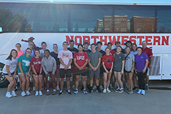 Members of the 2025-2026 Northwestern Oklahoma State University President's Leadership Class pose in two rows in front of white Northwestern charter bus with large red Northwestern lettering and Ranger mascot logo visible on bus exterior. Group of approximately 30 students and staff members wearing casual clothing including college t-shirts from various universities stands on concrete surface with brick Cunningham Hall building visible in background under sunny sky with rainbow. Tara Hannaford, Northwestern financial aid director, stands at right end of front row; Northwestern President Dr. Bo Hannaford stands at right end of back row with his son Tucker Hannaford; PLC mentor Maci Flowers stands at far right. Photo taken before July 2025 Dallas, Texas trip.