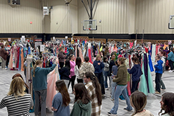 A large gymnasium filled with people browsing long rows of prom dresses organized on clothing racks. Signs on the racks indicate dress sizes. Multiple attendees of varying ages move through the space, some holding dresses. A basketball backboard is visible in the background. A