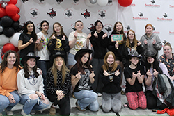 A group of approximately twelve young people pose in two rows in front of a step-and-repeat backdrop featuring the Northwestern Oklahoma State University and Social Work Department logos, flanked by red, black, and white balloon clusters. Several individuals in the front row hold prom dresses in dry-cleaning bags and prop signs with phrases including