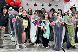 A group of approximately fifteen young people pose in two rows — some standing, some kneeling — in front of a step-and-repeat backdrop featuring the Northwestern Oklahoma State University and Social Work Department logos, with red, black, and white balloon clusters visible at the left. Several individuals make heart gestures with their fingers. One person holds a prop sign. One adult is visible at the far right.