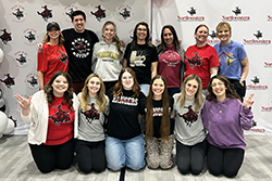 Thirteen volunteers pose in two rows — seven standing in the back row and six kneeling in the front — in front of a step-and-repeat backdrop featuring the Northwestern Oklahoma State University and Social Work Department logos, with white, black, and silver balloon clusters visible on each side. Most individuals wear Northwestern Rangers or Woodward-branded apparel in red, black, gray, and white. Back row, left to right: Dr. Kylene Rehder, Jonathan Wales, Kailie Martin (instructor of social work), Stefanie Alexander (Primp for Prom co-director), Melaine Yager, Katie Vaverka, Jennifer Pribble (instructor of social work). Front row, left to right: Krystin Pierce, Mattie Gore, Isabella Burden, Haylee Durrence, Kinley Rehder, and Codi Harding (assistant professor).