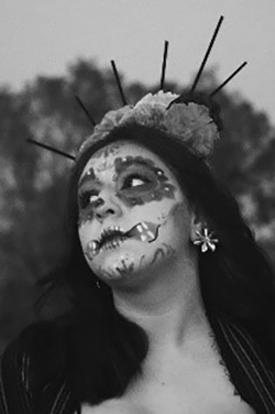 Black and white photograph of a person wearing Day of the Dead face paint with decorative skull designs, a flower crown, and ornamental hair accessories with vertical elements.