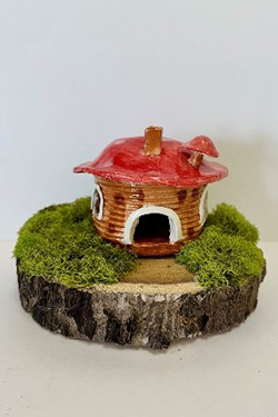 Ceramic sculpture of a small house with a red mushroom-shaped roof and chimney, set on a circular wood slice base covered with moss.