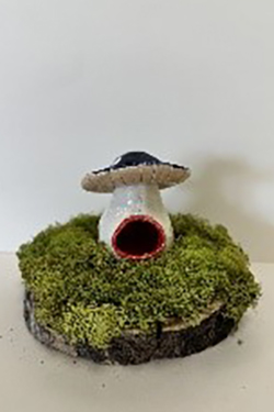 Ceramic sculpture of a mushroom-shaped house with a brown cap and cylindrical base with a rounded doorway, displayed on a circular wood slice base covered with moss.