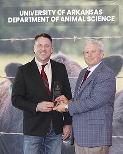 Dr. Dean Scarbrough, professor of agriculture and chair of the Agriculture Department at Northwestern Oklahoma State University, receives the 2026 University of Arkansas Department of Animal Science Advanced Graduate of Distinction Award. Scarbrough, wearing a black blazer and red tie, holds a glass award plaque while an unidentified presenter in a gray plaid blazer looks on. The pair stand before a University of Arkansas Department of Animal Science banner featuring a cattle photograph.
