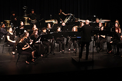 A large concert band performs on a darkened stage in Fellers Family Auditorium. Approximately 30 student musicians dressed in all-black attire are seated in a curved arrangement, playing woodwind, brass, and percussion instruments. Music stands with sheet music are positioned in front of each performer. A bald conductor in a black suit stands with his back to the camera, facing the ensemble on a low riser. Various instruments visible include clarinets, flutes, trombones, a tuba, and a bass drum. Stage lighting illuminates the performers against a dark background.