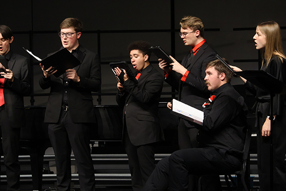Six members of the Northwestern Oklahoma State University University Singers perform on stage during the Holiday Gala in December 2025. The singers wear black formal attire with red ties and accessories and hold black music folders and a tablet. They are actively singing, with mouths open and engaged expressions, against a dark stage backdrop with tiered risers visible behind them.