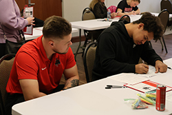 Northwestern Oklahoma State University admissions representative in red Nike polo assists prospective student in black Nike hoodie completing application paperwork at white table with promotional materials and pens during Spring Showcase event in Student Center conference room with multiple other students visible working on applications in background.