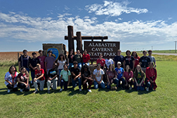 Approximately 30 high school students wearing casual summer clothes and red lanyards pose in two rows in front of Alabaster Caverns State Park entrance sign with rustic wooden posts, green lawn foreground, and blue sky with white clouds over plowed field and grassland in background.