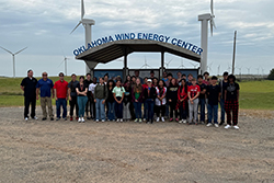 Group of approximately 35 high school students and adult staff members standing together under covered structure with Oklahoma Wind Energy Center sign overhead, multiple white wind turbines visible in grassy field behind group under cloudy sky