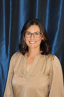 Morgan Dutton, Northwestern Oklahoma State University's Spring 2026 'Teach Like a Ranger' honoree, smiles in a professional portrait. She has shoulder-length dark hair, wears rectangular-framed glasses and a champagne-colored blouse, and is photographed against a draped blue fabric backdrop.