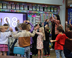 Cecely Franz and paraprofessional Starlet James dance with kindergarten students during classroom activity at Washington Early Childhood Center. Large number display on whiteboard shows 98, with students participating actively in movement-based learning activity.