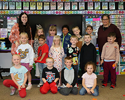 Cecely Franz, Northwestern Oklahoma State University alumna and kindergarten teacher, poses with her kindergarten class and paraprofessional Starlet James in classroom at Washington Early Childhood Center in Alva, Oklahoma. Students arranged in two rows with alphabet wall display and colorful smiley face decorations visible in background.