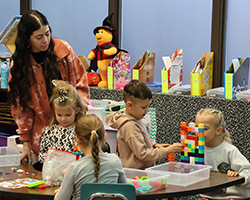 Kindergarten teacher Cecely Franz interacts with students during hands-on learning activity with building blocks at Washington Early Childhood Center. Students engaged in collaborative play while teacher provides guidance at classroom table.