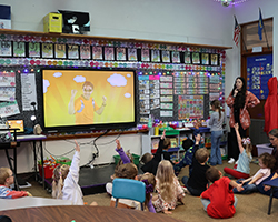 Northwestern alumna Cecely Franz answers student question during kindergarten lesson at Washington Early Childhood Center in Alva, Oklahoma. Students seated on carpet with hands raised, alphabet learning materials and classroom calendar visible on walls.