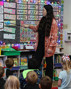 Kindergarten teacher Cecely Franz leads morning classroom activities with students at Washington Early Childhood Center. Students raise hands enthusiastically while seated on carpet, with interactive whiteboard displaying educational content and calendar board visible on classroom wall.