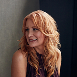 Dr. Constance Squires, author and creative writing professor at the University of Central Oklahoma, photographed in a professional portrait. She has long, wavy auburn hair and is wearing a sleeveless dark purple top. She is smiling and looking slightly off-camera to her left, posed against a blurred two-tone background of gray and dark blue.