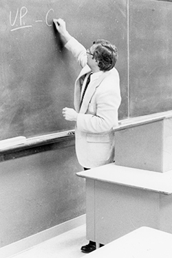 Black and white archival photograph showing a person in a light-colored blazer and dark pants writing 