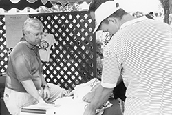 Black and white archival photograph showing two people at an outdoor event with a lattice fence backdrop. One person wearing glasses and a t-shirt with a graphic design stands on the left near a table with a price list sign. Another person wearing a white cowboy hat and striped polo shirt stands on the right, appearing to interact at the table. A festive scalloped awning is visible above.