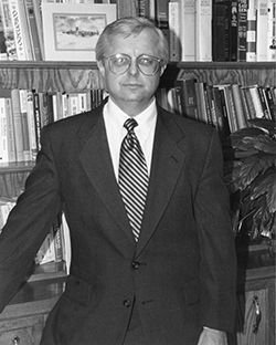 Black and white photograph of a person wearing a dark suit jacket, white dress shirt, and diagonal striped tie, standing in an office with bookshelves. The person wears large-framed glasses and stands in front of floor-to-ceiling bookshelves filled with books. A framed picture is visible on the upper shelf, and a potted plant appears on the right side of the frame.