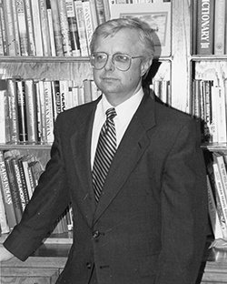 Professional headshot photograph of Dr. Timothy Zwink wearing a dark suit jacket, white dress shirt, and patterned tie, standing in front of a bookshelf filled with books. The person wears glasses and has light-colored hair.