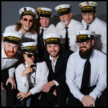 Eight members of King Cabbage Brass Band posed together against a gray studio backdrop, all wearing matching white nautical-style caps labeled 
