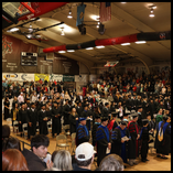 Wide-angle photograph of Northwestern Oklahoma State University's Fall Commencement ceremony in Percefull Fieldhouse. Graduates in black caps and gowns are seated in rows in the center of the gymnasium floor, with faculty in colorful academic regalia seat
