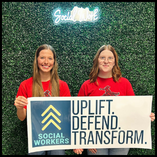 Two young women in red Northwestern Oklahoma State University Ranger t-shirts stand smiling in front of a green hedge wall with a neon 