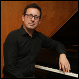 Dr. Eduardo Moreira, Northwestern Oklahoma State University assistant professor of music, seated at black grand piano wearing dark button-down shirt and glasses, positioned for professional portrait in warm wood-paneled room.