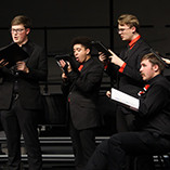 Six members of the Northwestern Oklahoma State University University Singers perform on stage during the Holiday Gala in December 2025. The singers wear black formal attire with red ties and accessories and hold black music folders and a tablet. 