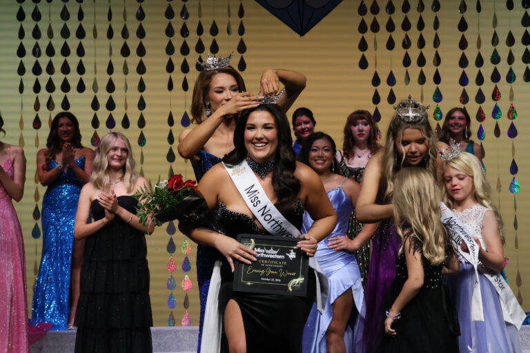 Abbey Elam, Miss Northwestern 2026, in a royal blue sequined gown and crown, places the Miss Northwestern crown on a smiling Raegan Bleything, who wears a black gown and Miss Northwestern sash and holds red roses and her evening gown award certificate, as contestants applaud on stage in Fellers Family Auditorium.