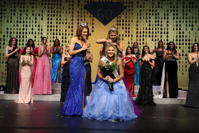 Lauren Thomason, Miss Northwestern’s Teen 2026, in a purple gown, places the Teen crown on a smiling Nelley Burnett, who kneels in a light blue ball gown with a Miss Northwestern’s Teen sash and a bouquet, as Miss Northwestern 2026 Abbey Elam, in royal blue, holds her hands to her heart on stage in Fellers Family Auditorium.