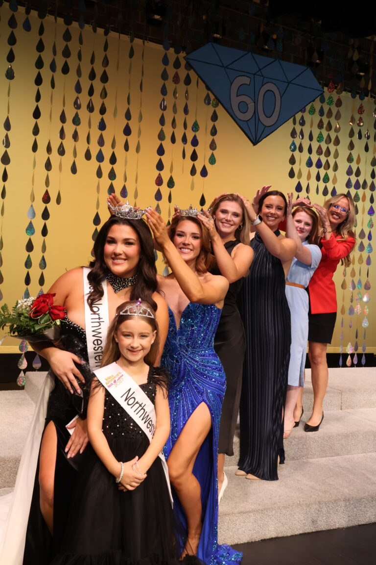 Five former Miss Northwesterns stand in a line on the steps behind Miss Northwestern 2027 Raegan Bleything, each holding her hands above the head of the woman in front of her like a crown, on stage in Fellers Family Auditorium. Bleything wears a black gown, crown and Miss Northwestern sash and holds red roses. Northwestern Star 2027 Harper Higgins, in a black dress, tiara and Northwestern Star sash, stands in front of her. A blue diamond marked 60 hangs on the gold backdrop above them.